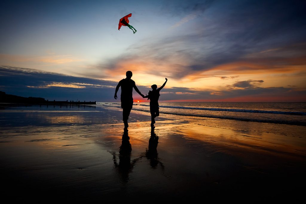 A couple hand in hand flying a kite at sunset along Cromer Beach at low tide in silhouette.