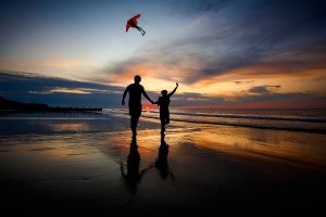 A couple hand in hand flying a kite at sunset along Cromer Beach at low tide in silhouette.