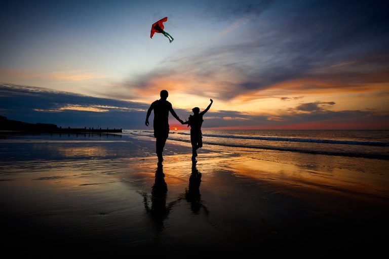A couple hand in hand flying a kite at sunset along Cromer Beach at low tide in silhouette.